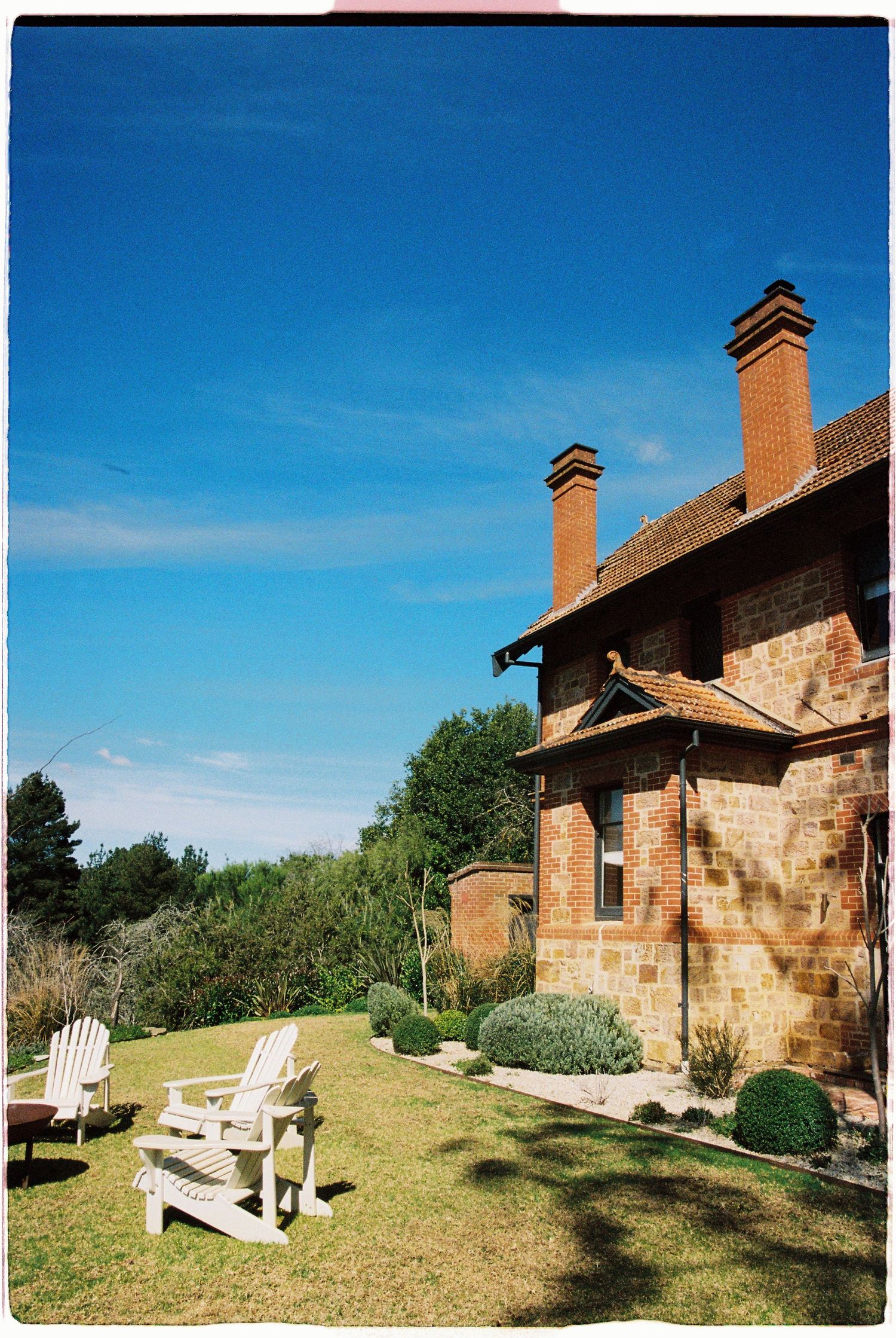 Rustic brick country house with two chimneys, white Adirondack chairs on green lawn under blue sky.