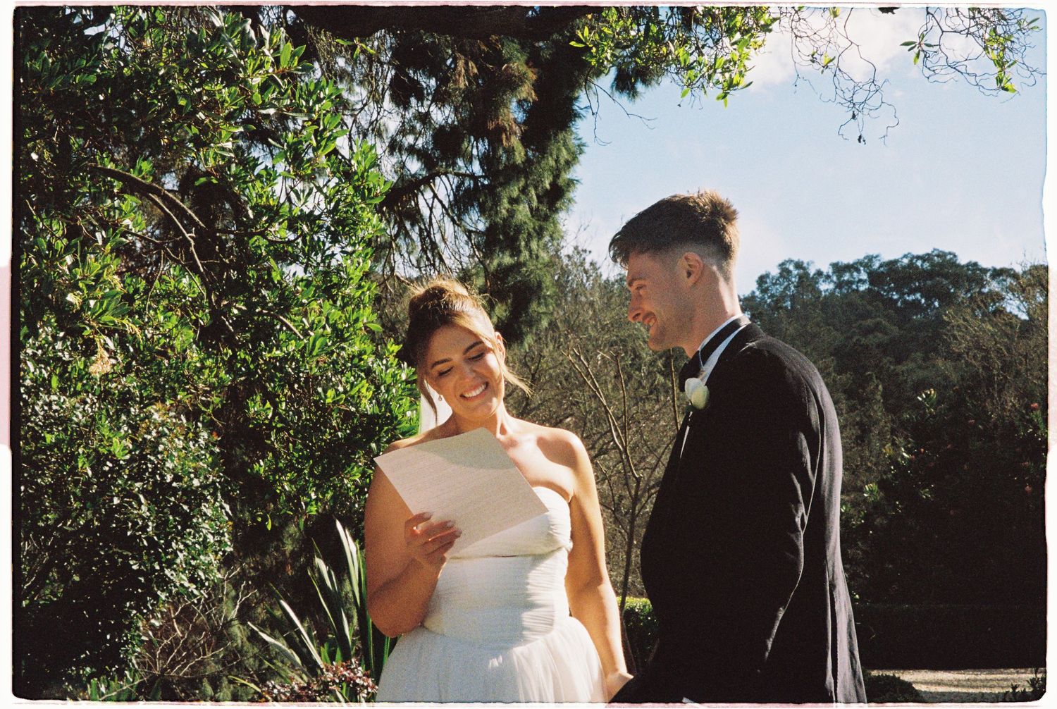 Bride reading vows to groom during outdoor wedding ceremony surrounded by greenery.