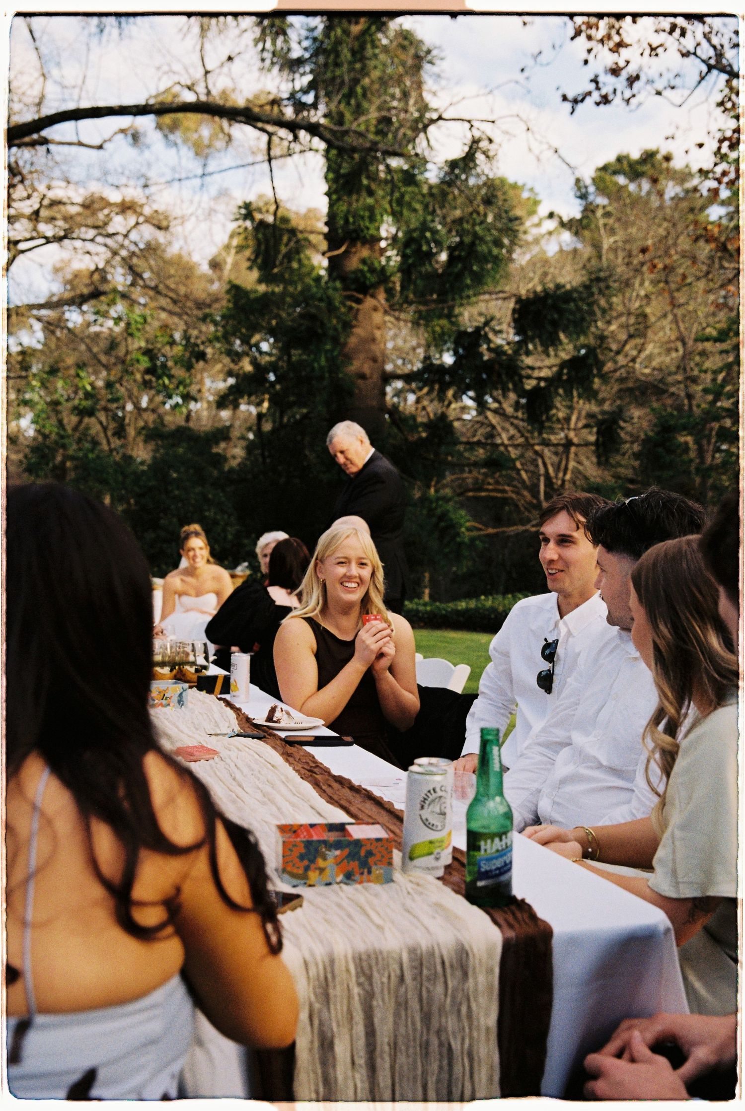 Guests laughing and socializing at an outdoor wedding reception table surrounded by lush green trees.