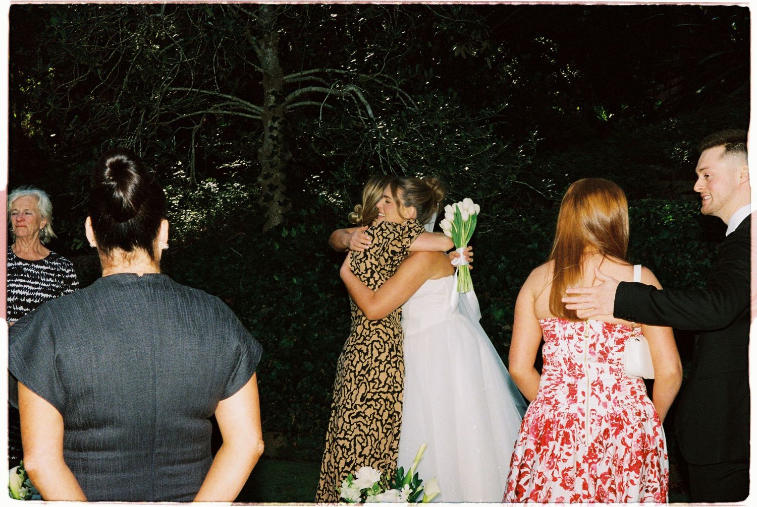 Wedding guests embracing the bride in a white gown during an outdoor garden ceremony at dusk.