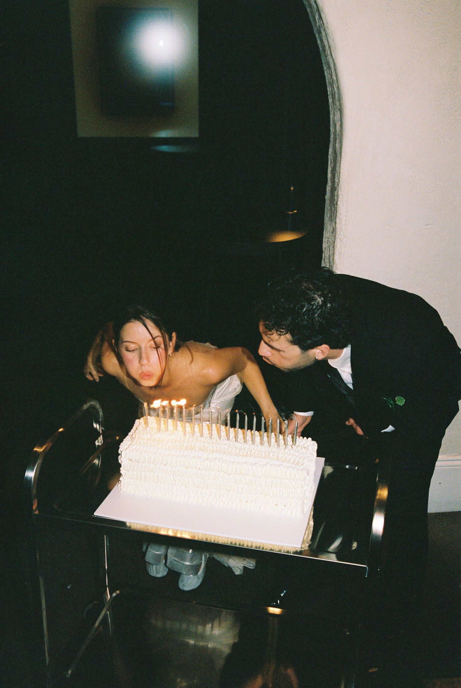 Bride and groom blowing out candles on a white tiered wedding cake at a reception.