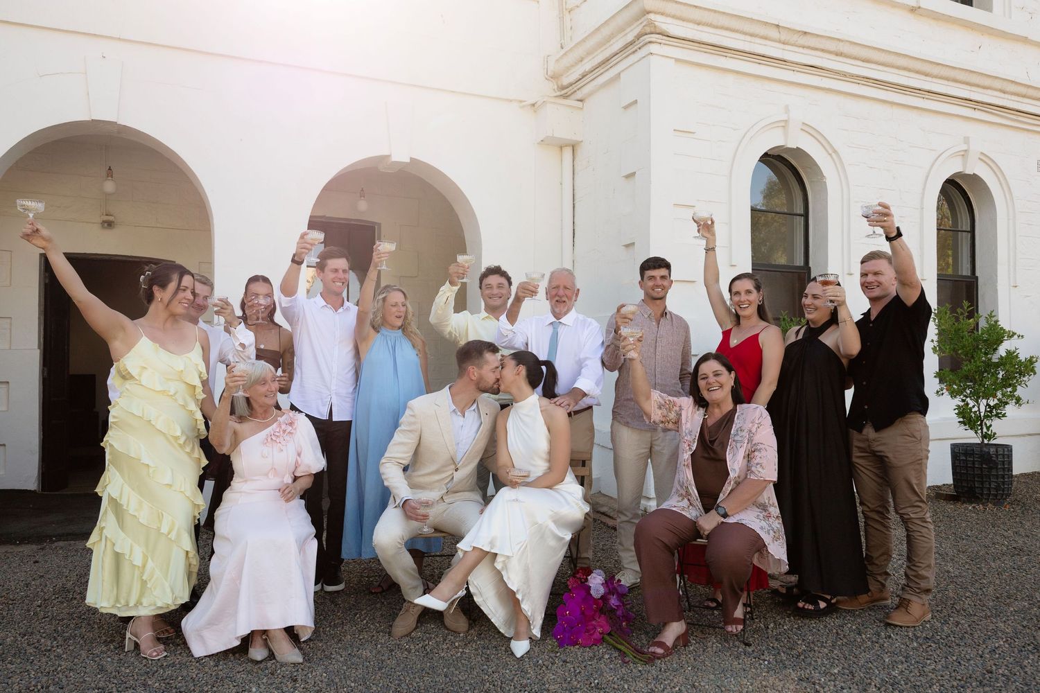 Wedding group photo with guests raising glasses in toast outside elegant white building.