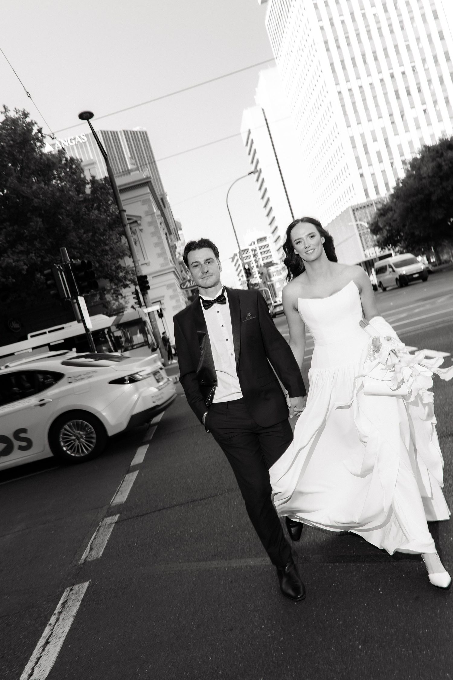 Bride and groom walk boldly across a city street in black and white wedding attire with urban buildings behind.