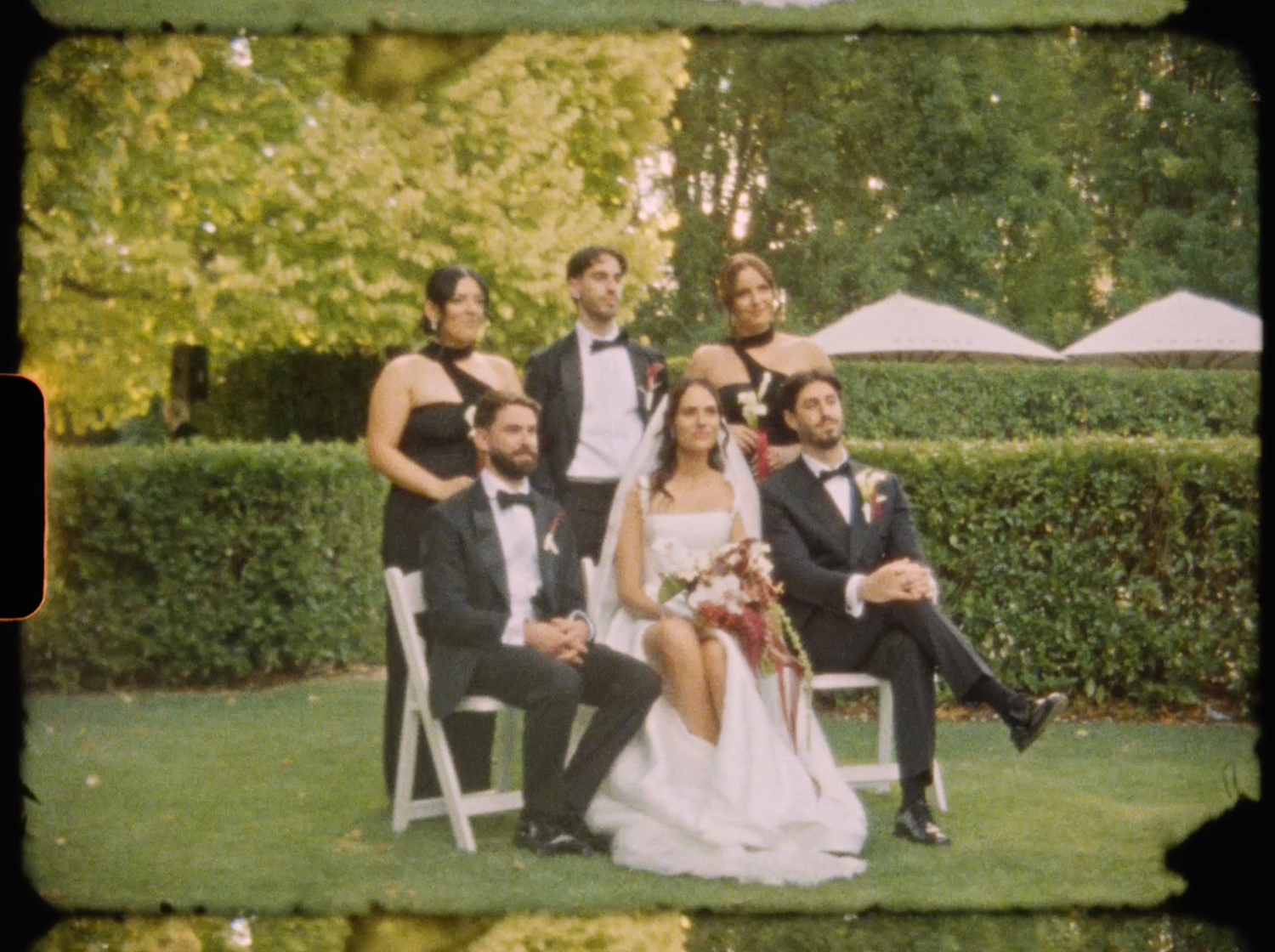 Bride and groom pose with wedding party on manicured garden lawn in elegant formal wedding attire.