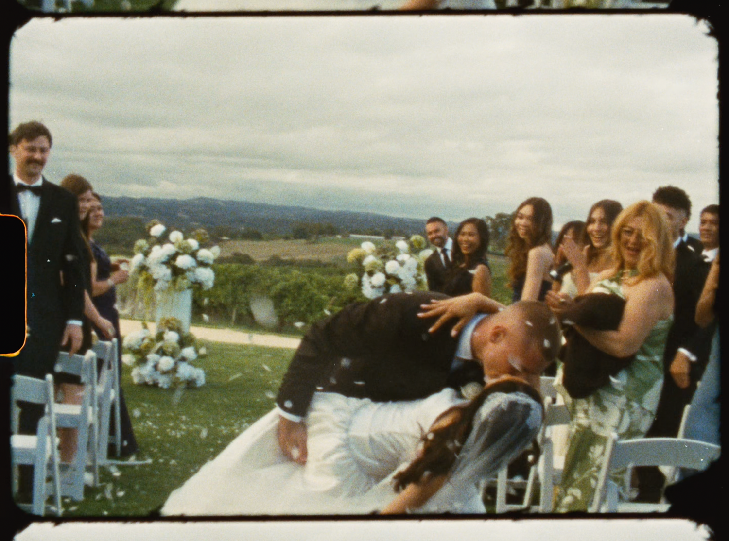 Groom dramatically dipping bride in white gown during outdoor wedding celebration with guests watching.