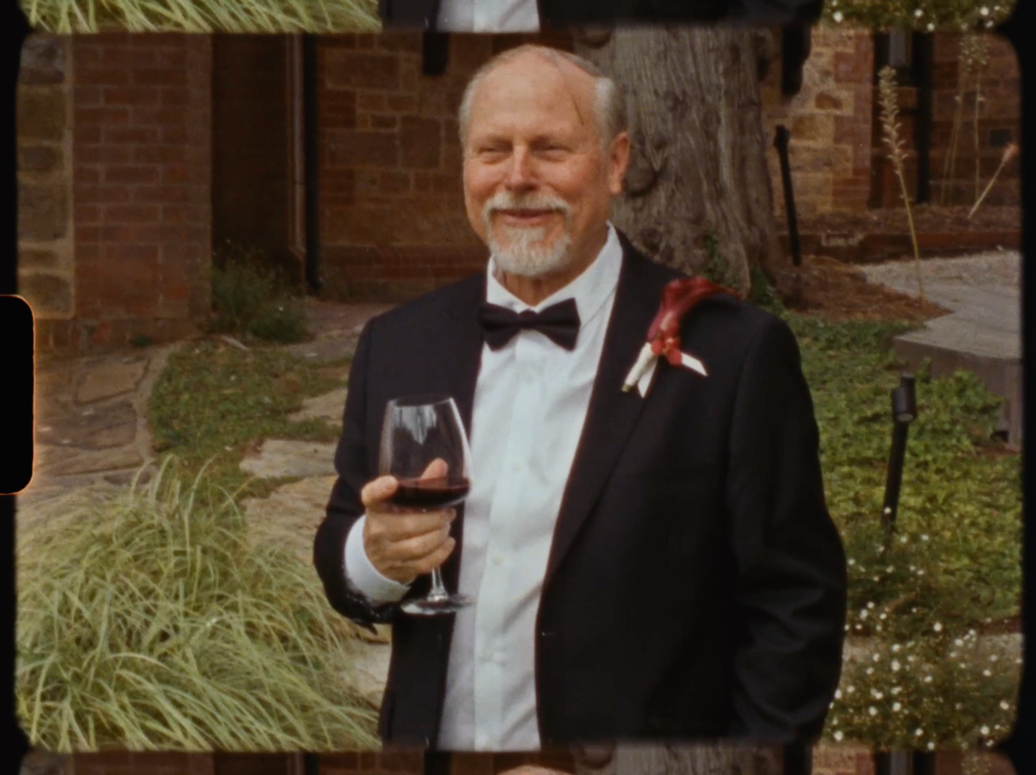 Smiling older man in tuxedo with bow tie and boutonniere holding red wine glass at wedding.