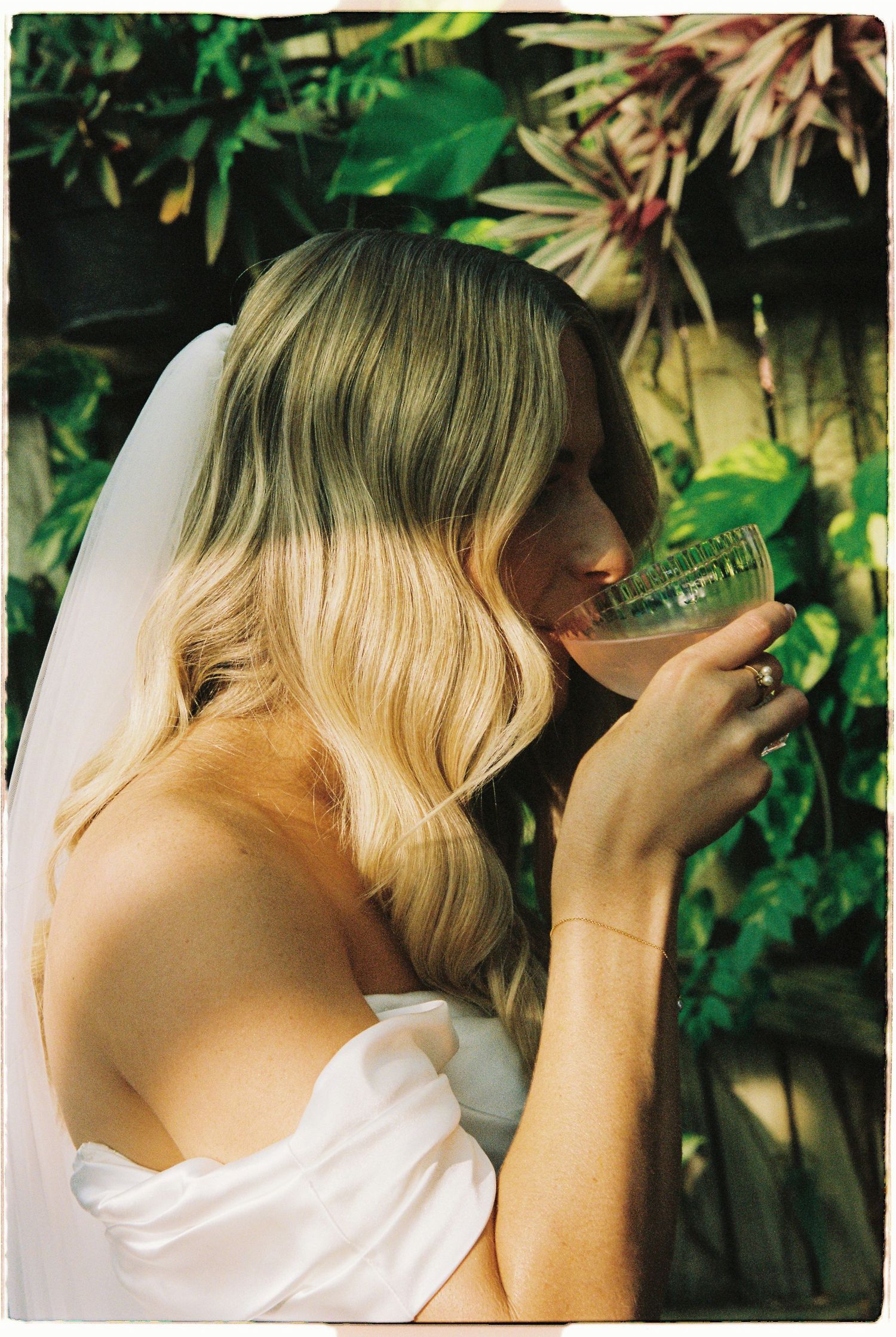 Blonde bride in white strapless gown sips a cocktail surrounded by tropical greenery.