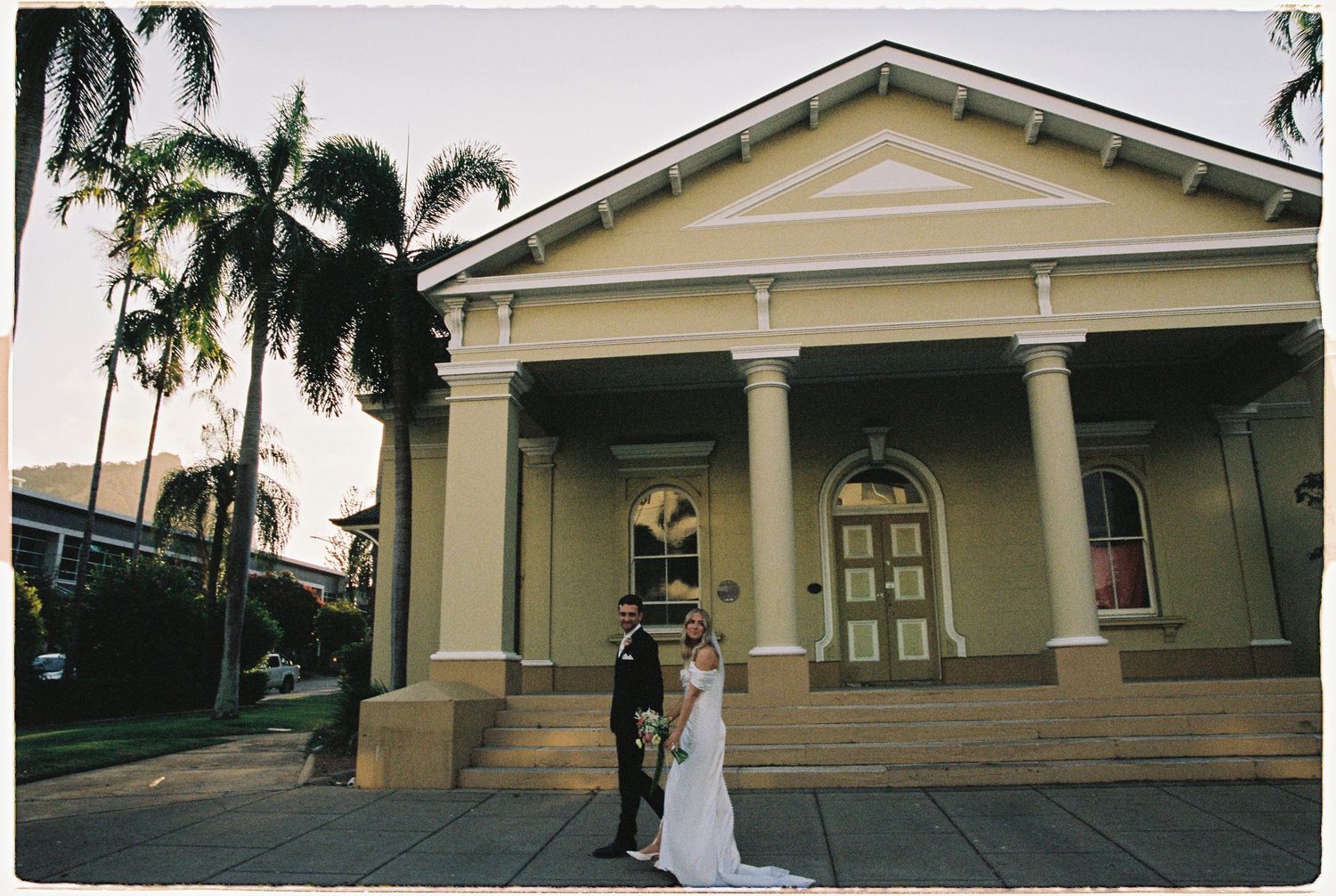 Wedding couple poses outside elegant colonial building with palm trees at sunset.