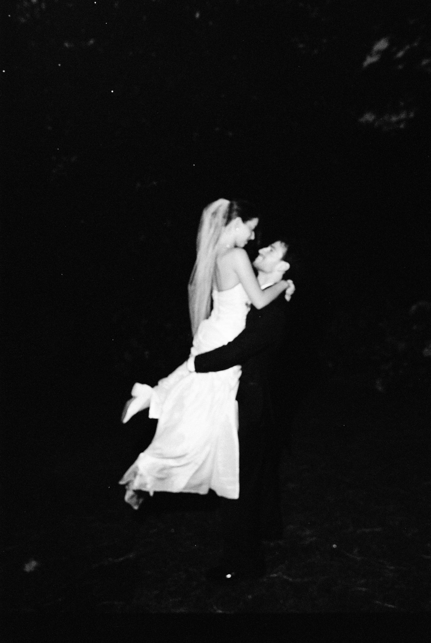Black and white film photo of groom lifting bride in wedding dress during a romantic first dance.