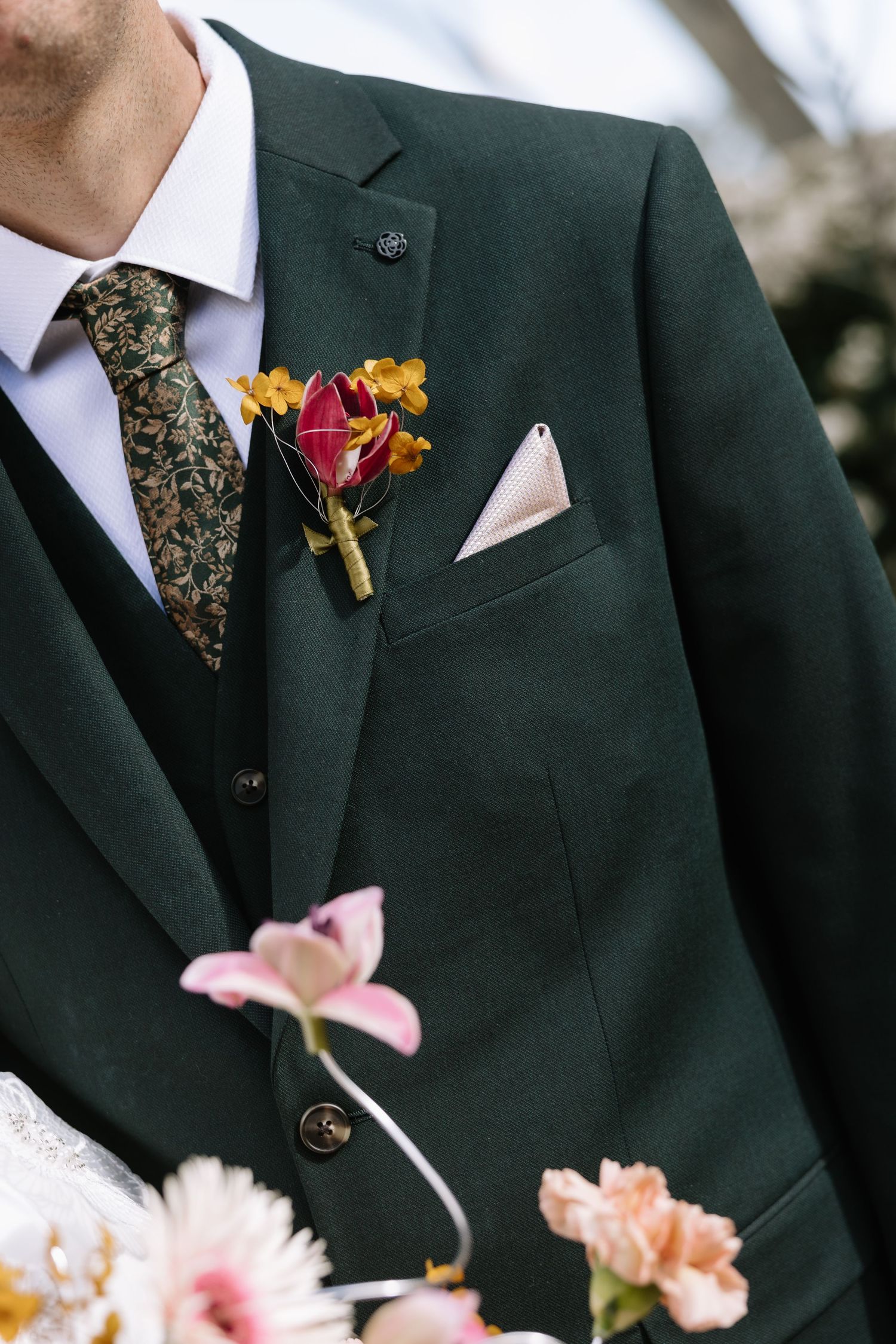 Groom in dark green suit with floral tie, boutonniere, and white pocket square, surrounded by colorful wedding flowers.