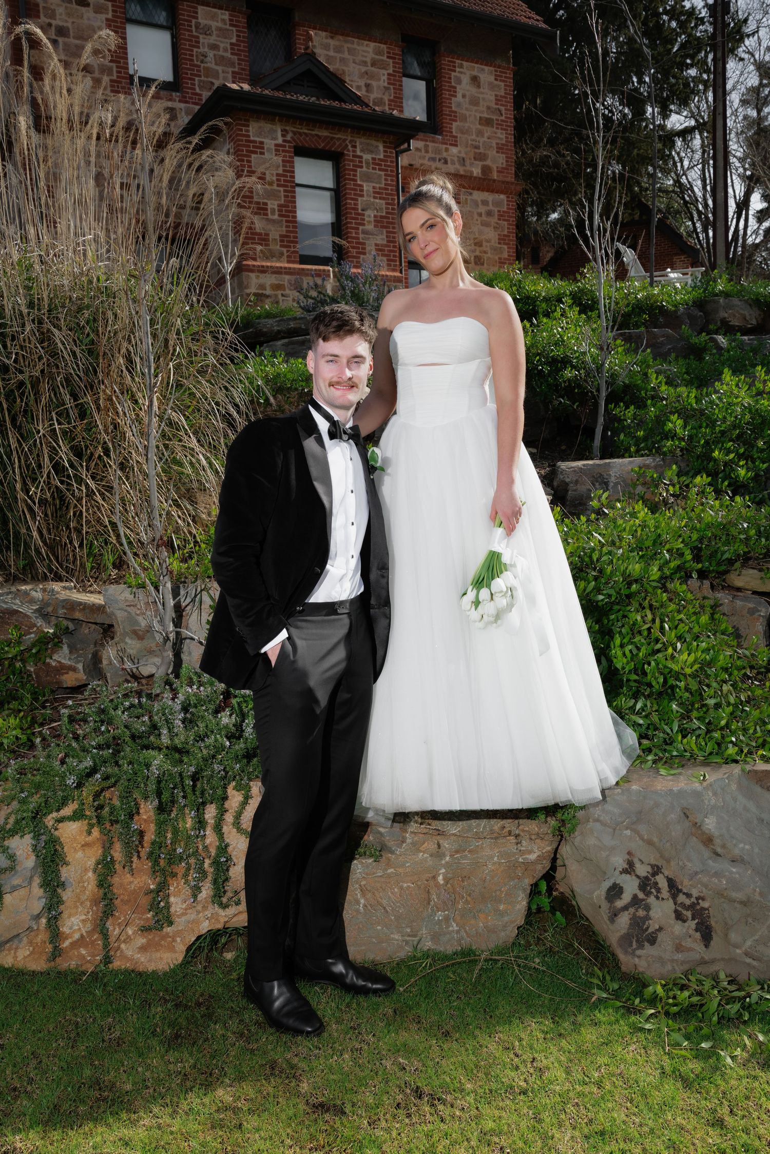 Bride in white strapless gown holding bouquet stands on rock beside groom in black tuxedo at outdoor wedding venue.