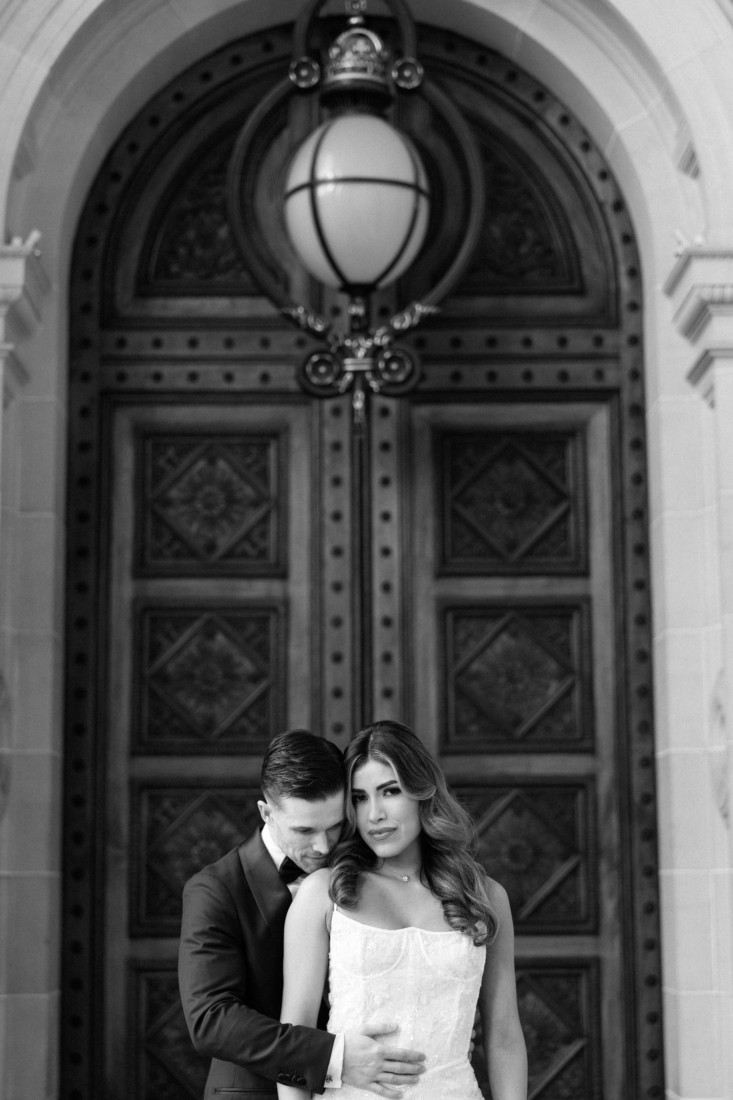 Black and white photo of bride and groom embracing before ornate wooden doors.