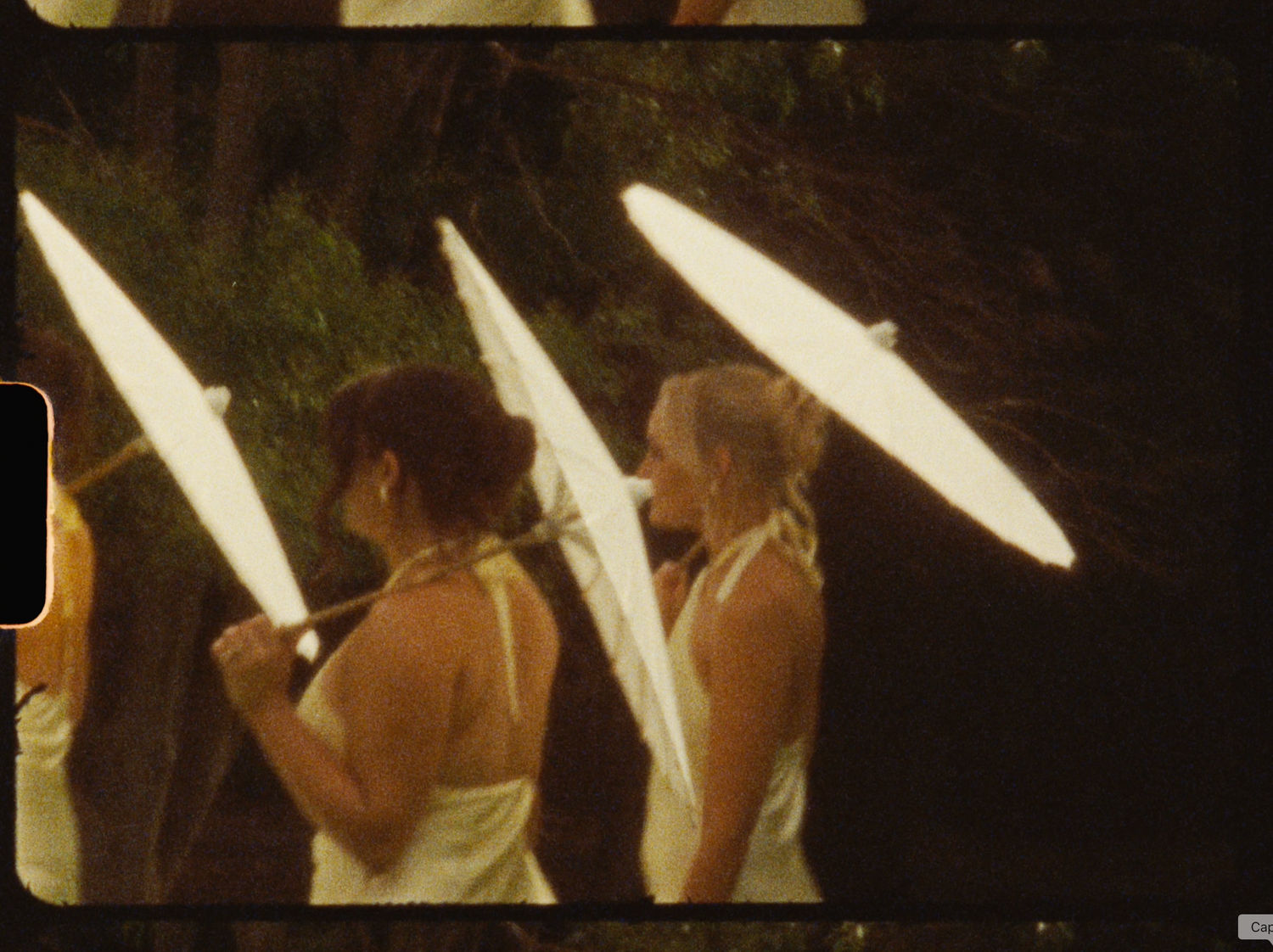 Two women in white dresses holding white parasols outdoors in a vintage film photograph.