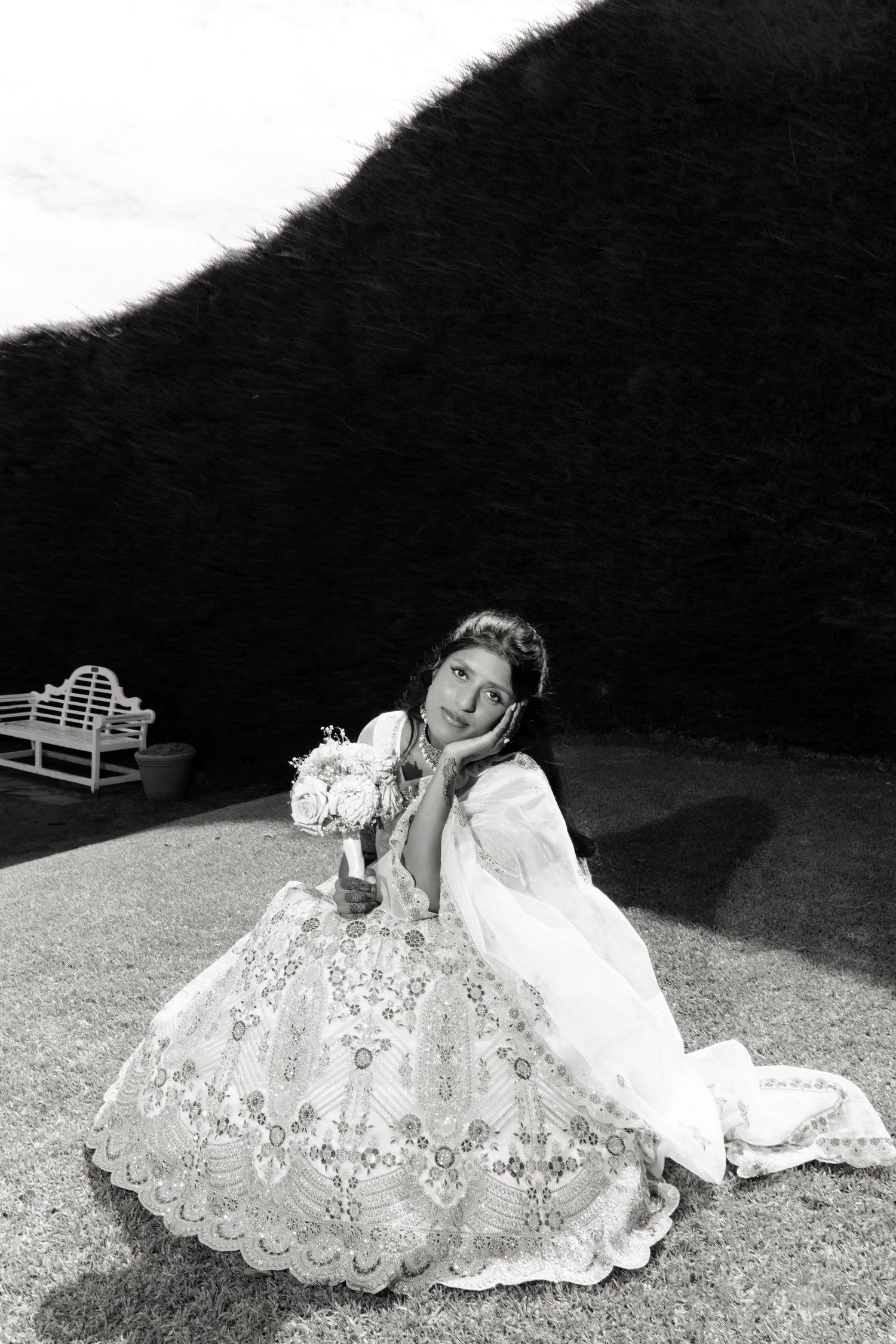Black and white photo of a bride in an elegant lace gown sitting on grass, holding a bouquet, with a bench in the background.