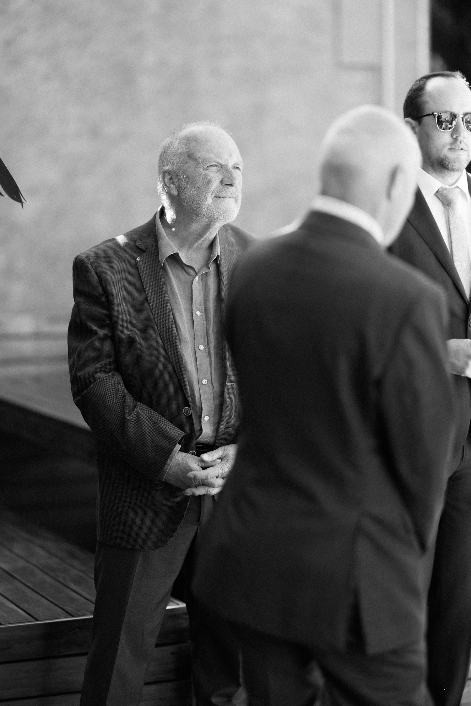 Black and white candid photo of elderly male wedding guest smiling during an outdoor ceremony.