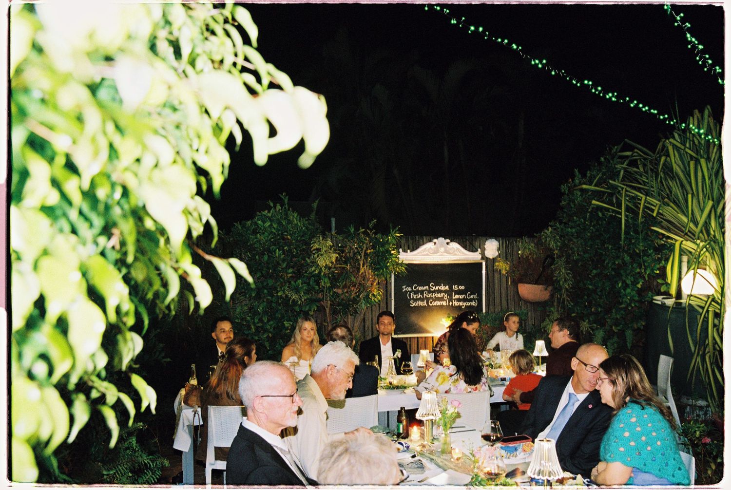 Guests dining outdoors at night under string lights surrounded by lush greenery at an elegant garden dinner party.
