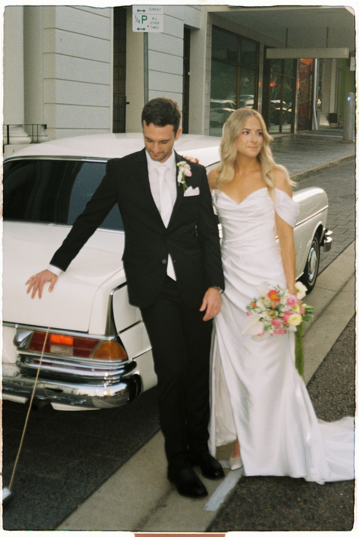 Bride in white gown holding floral bouquet stands with groom in black tuxedo beside a classic white car on a city street.