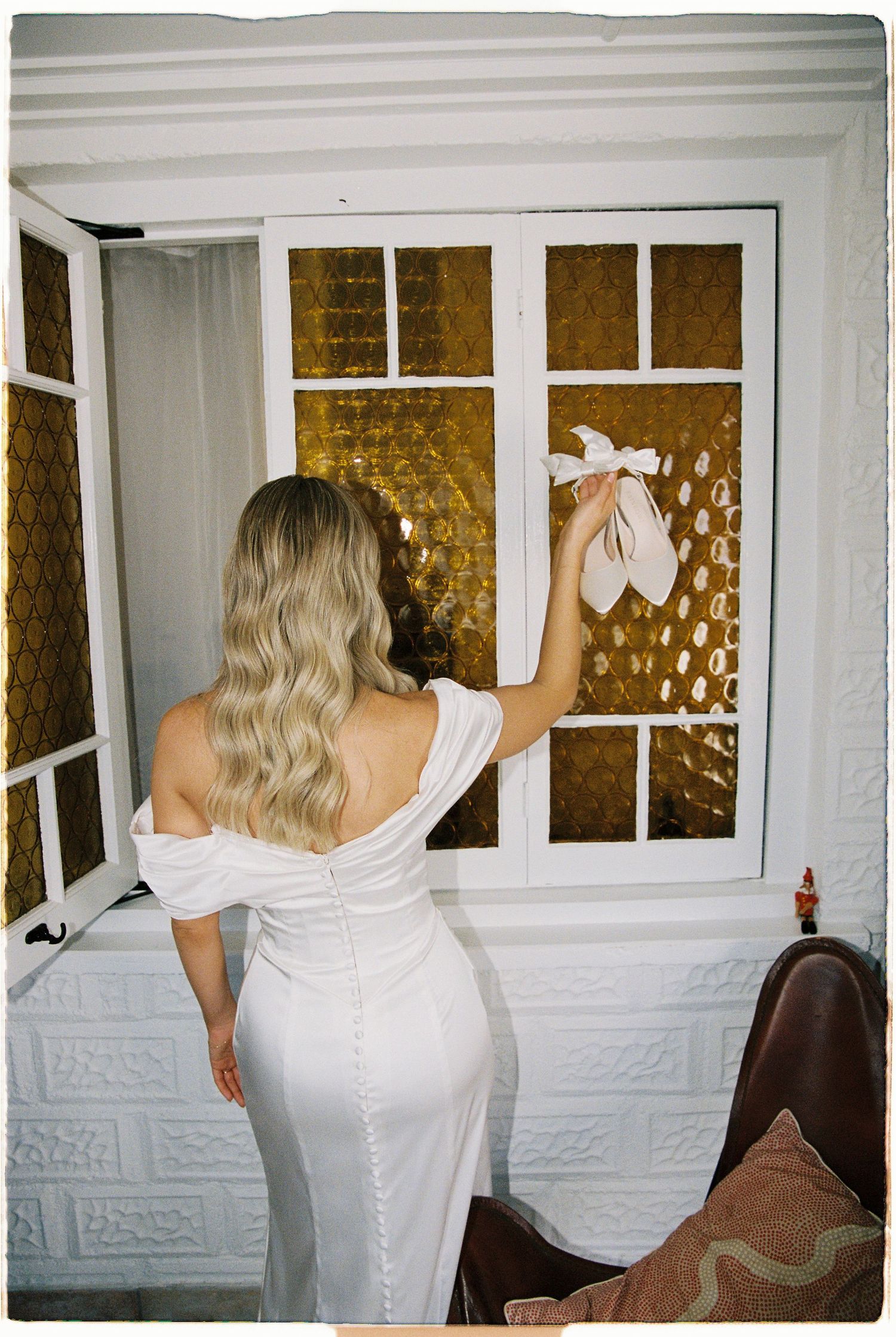 Bride in white off-shoulder gown holding heels with bow ribbon near golden stained glass window.