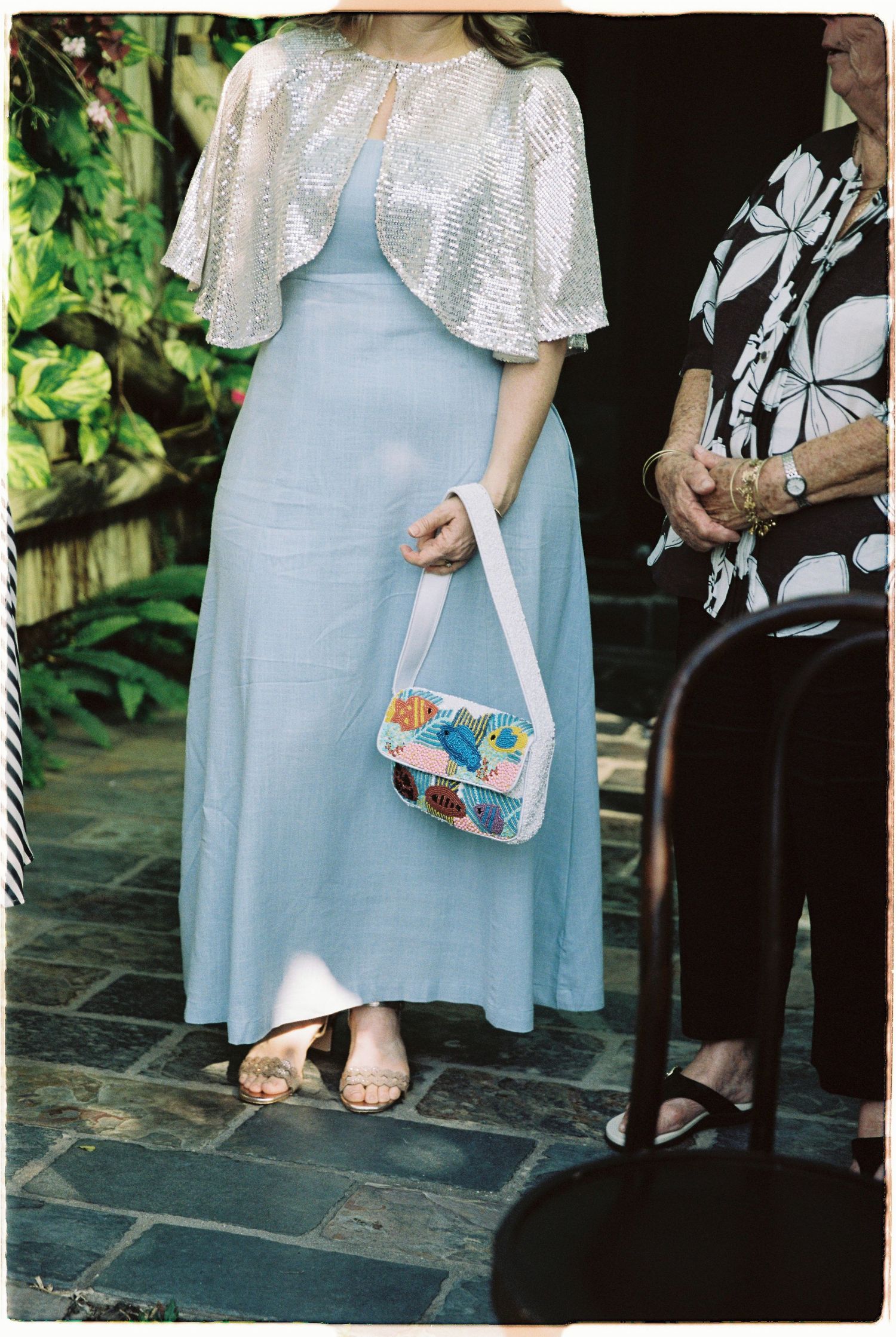Wedding guest in pale blue maxi dress with sequin cape holding colorful floral handbag.