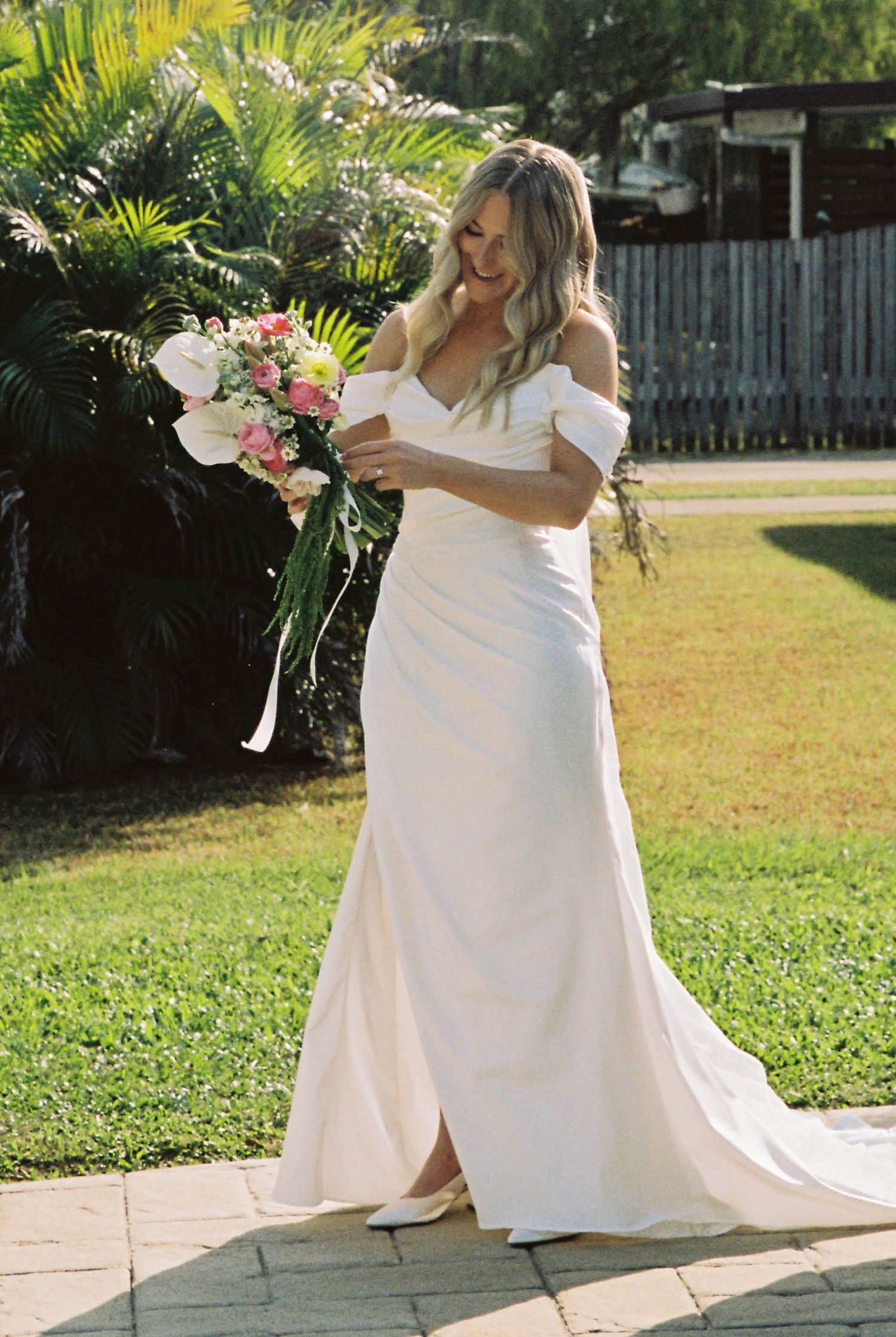 Smiling bride in off-shoulder white gown holding pink and white bouquet outdoors on sunny day.