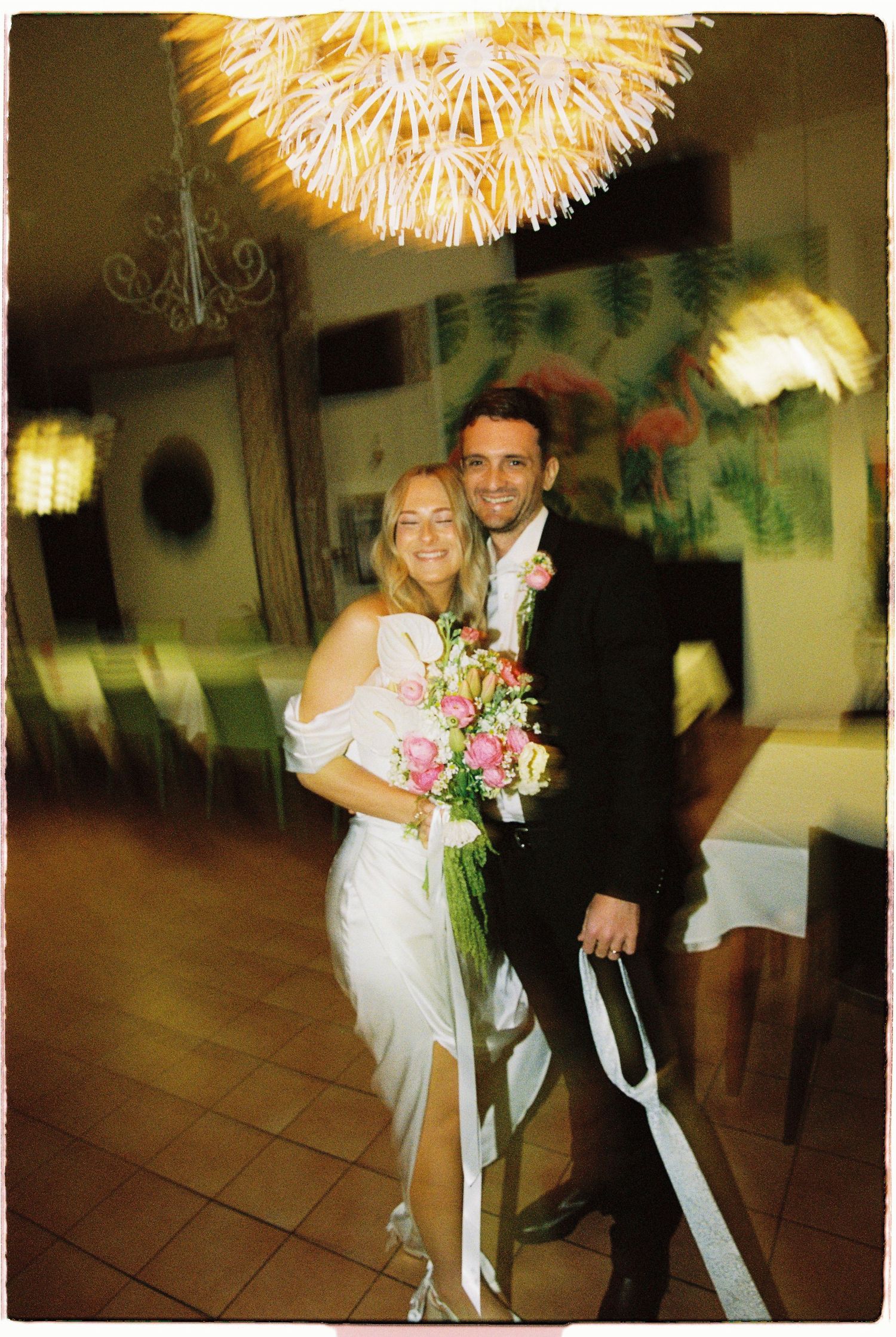 Happy newlywed couple posing together indoors under chandelier, bride holding pink and white bouquet.