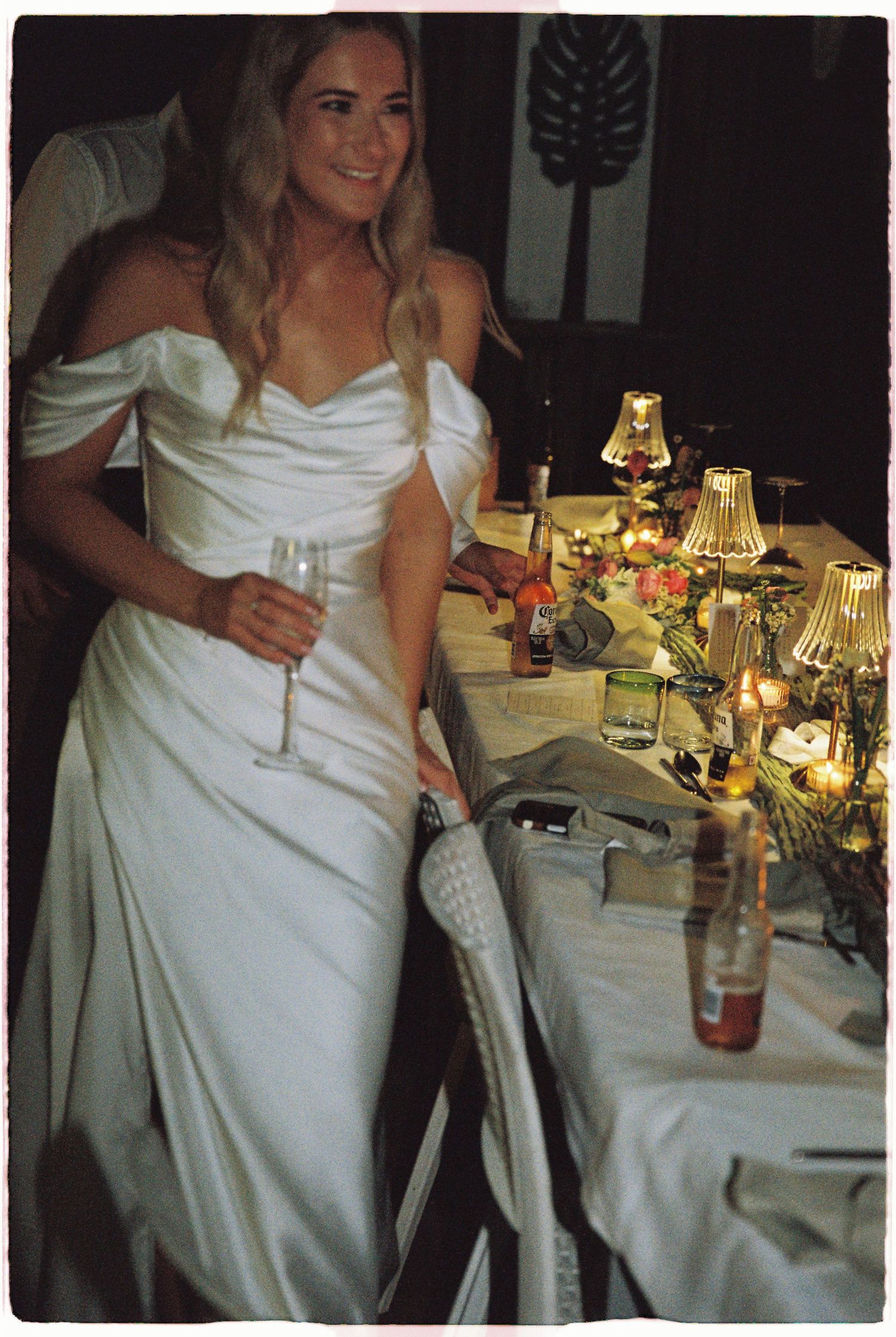A radiant bride in a white off-shoulder gown holds a champagne glass at a candlelit wedding reception table.