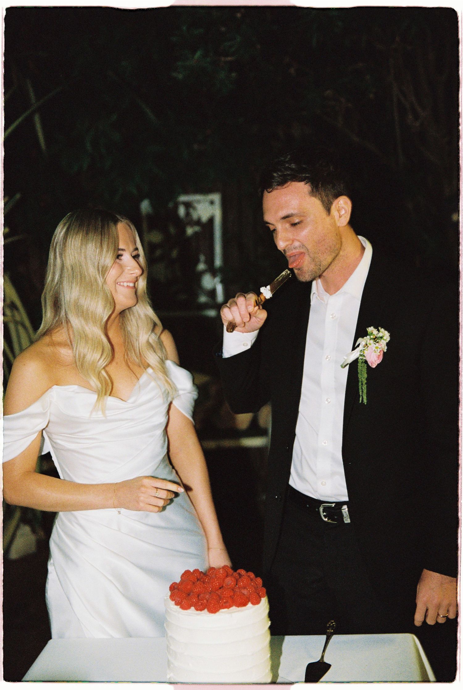 Bride and groom cutting a white wedding cake topped with fresh strawberries at their outdoor evening reception.