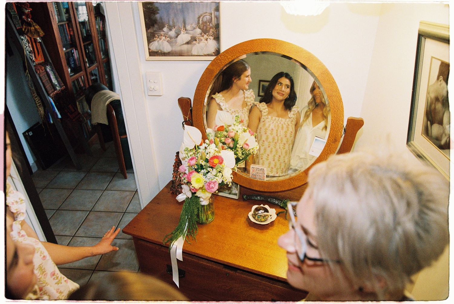 Bride admiring herself in round wooden vanity mirror surrounded by family before wedding.