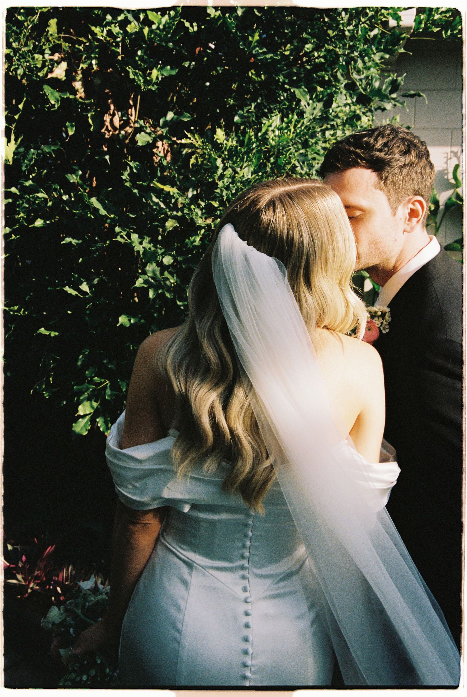 Bride and groom sharing a kiss in front of lush green garden foliage after ceremony.