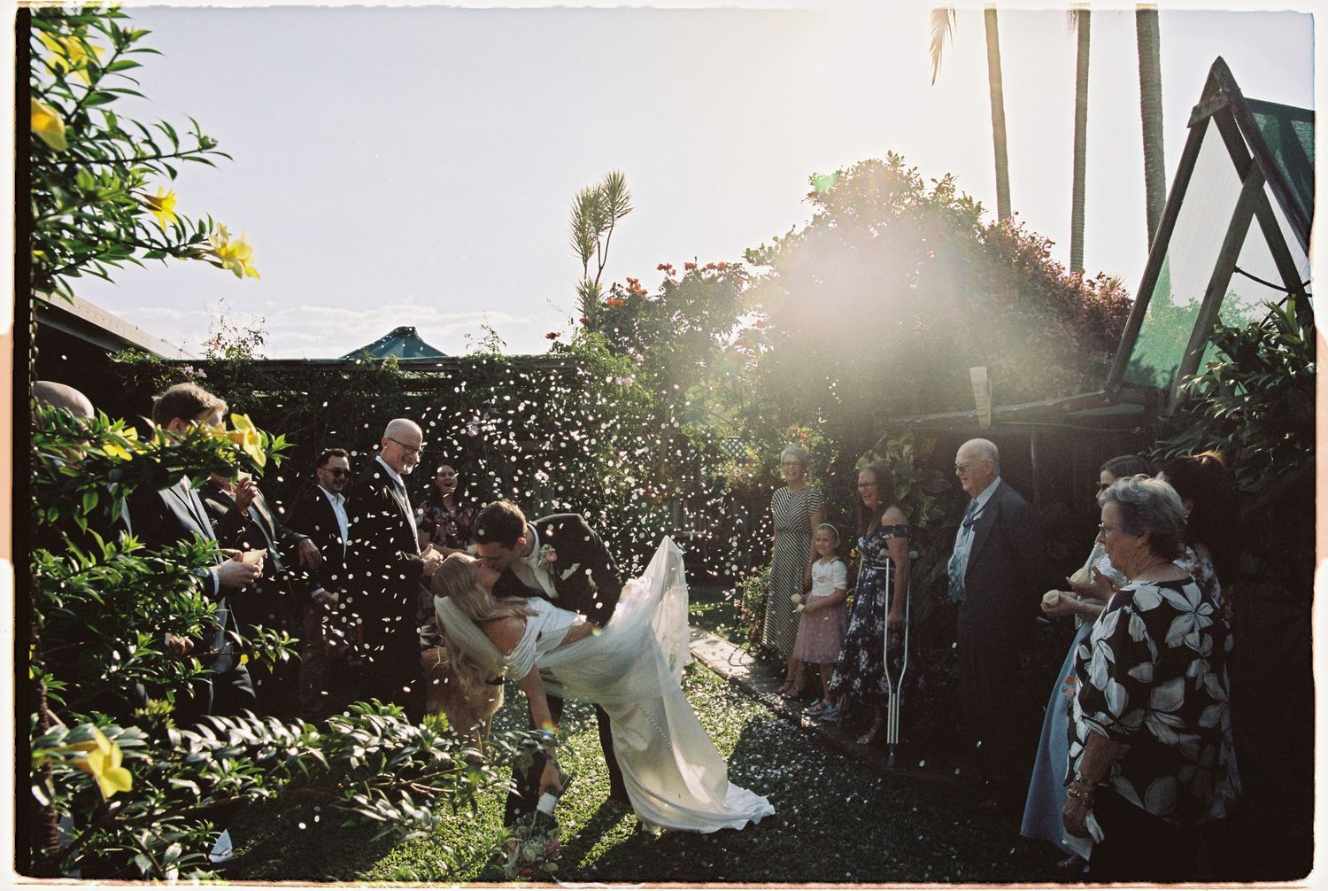 Groom lifts bride in white gown as guests shower couple with confetti in sunny garden.