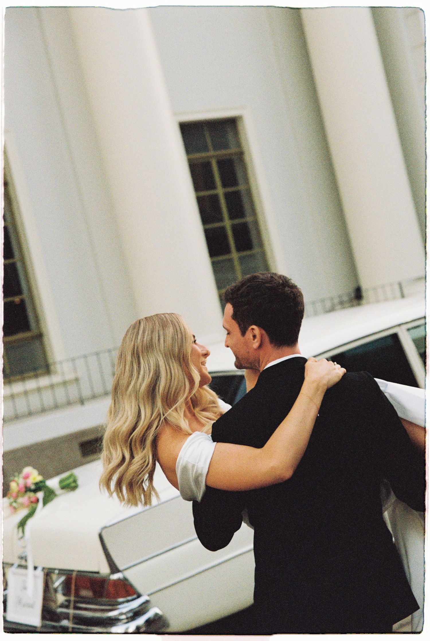 Bride and groom embracing romantically beside a vintage white wedding car outside a grand columned building.