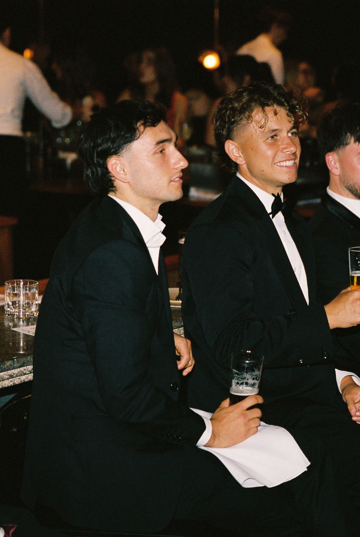 Two young men in black tuxedos smile and watch wedding speeches at a formal reception dinner.