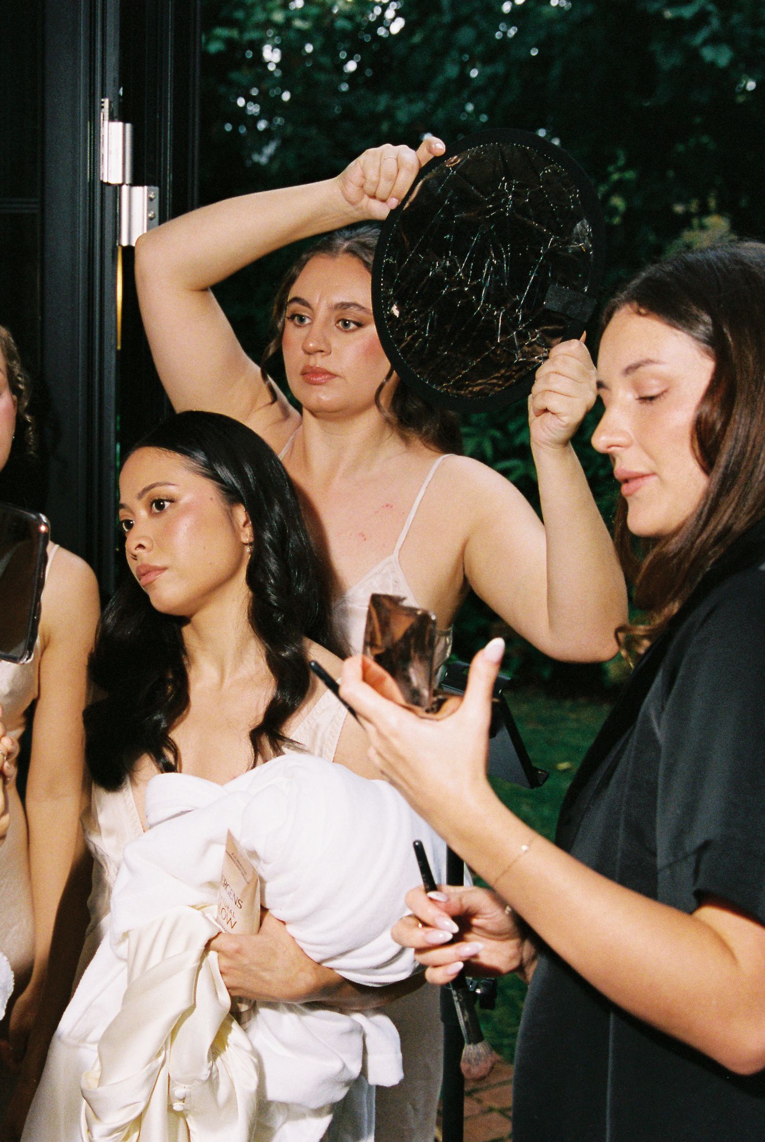 Bridesmaids in silk slips touching up makeup with decorative mirror outdoors before ceremony.