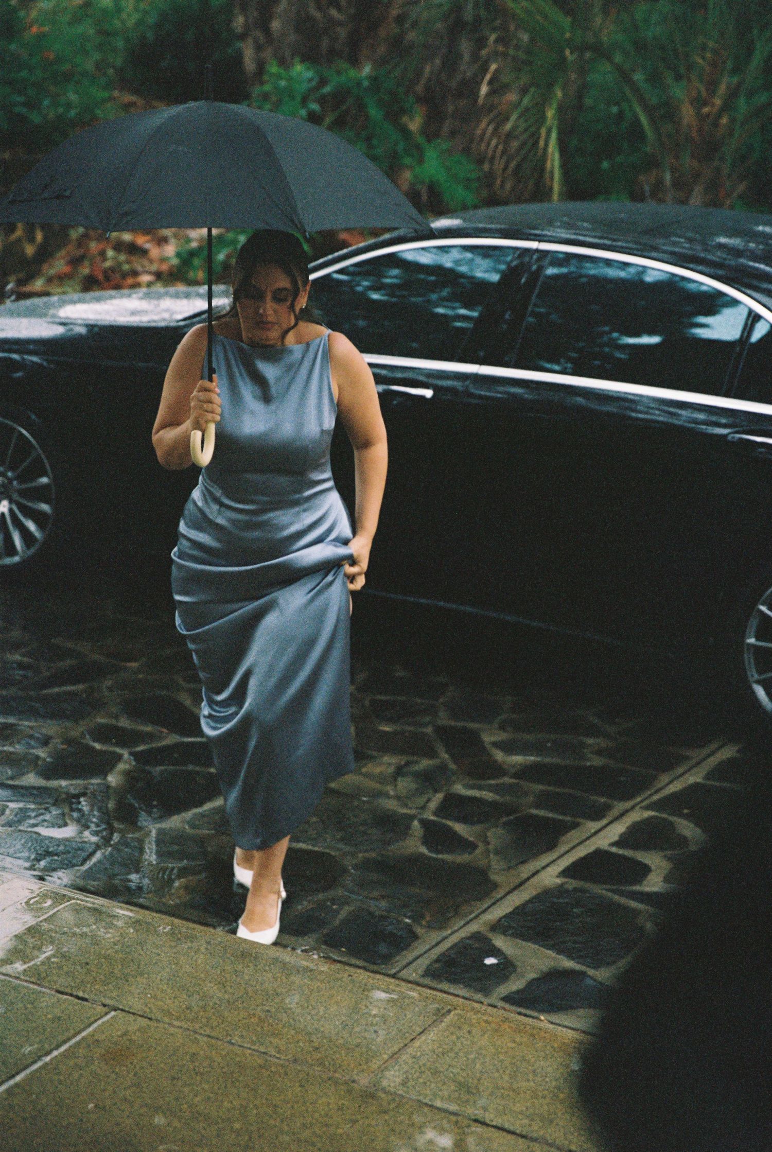 Woman in silver satin dress holding black umbrella walking in rain at wedding venue.