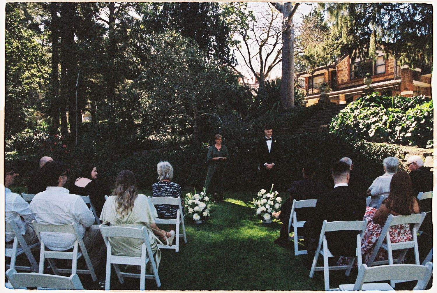 Intimate outdoor garden wedding ceremony with white chairs and lush green trees at a historic estate.