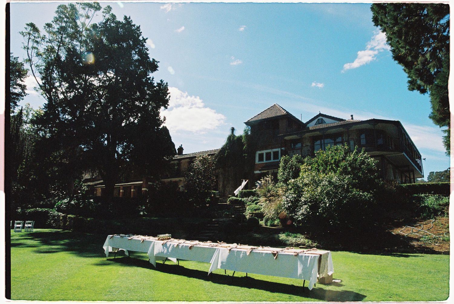 Elegant outdoor wedding reception with long white draped table set on manicured lawn before a grand estate manor.