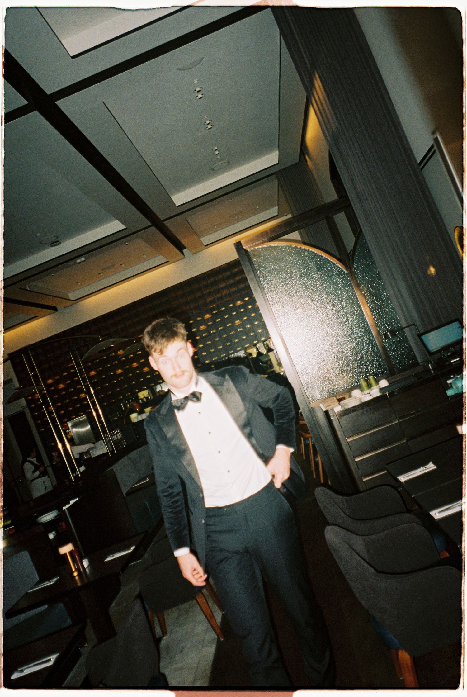 Groom in black tuxedo and bow tie poses confidently in an elegant dark restaurant interior.