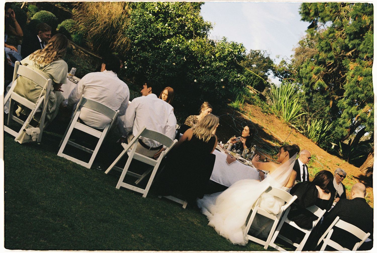 Aerial view of wedding guests seated at outdoor reception tables on sloped garden lawn.