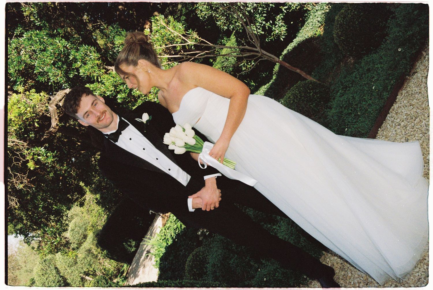 Bride and groom posing together in garden, she in strapless white gown holding white roses.