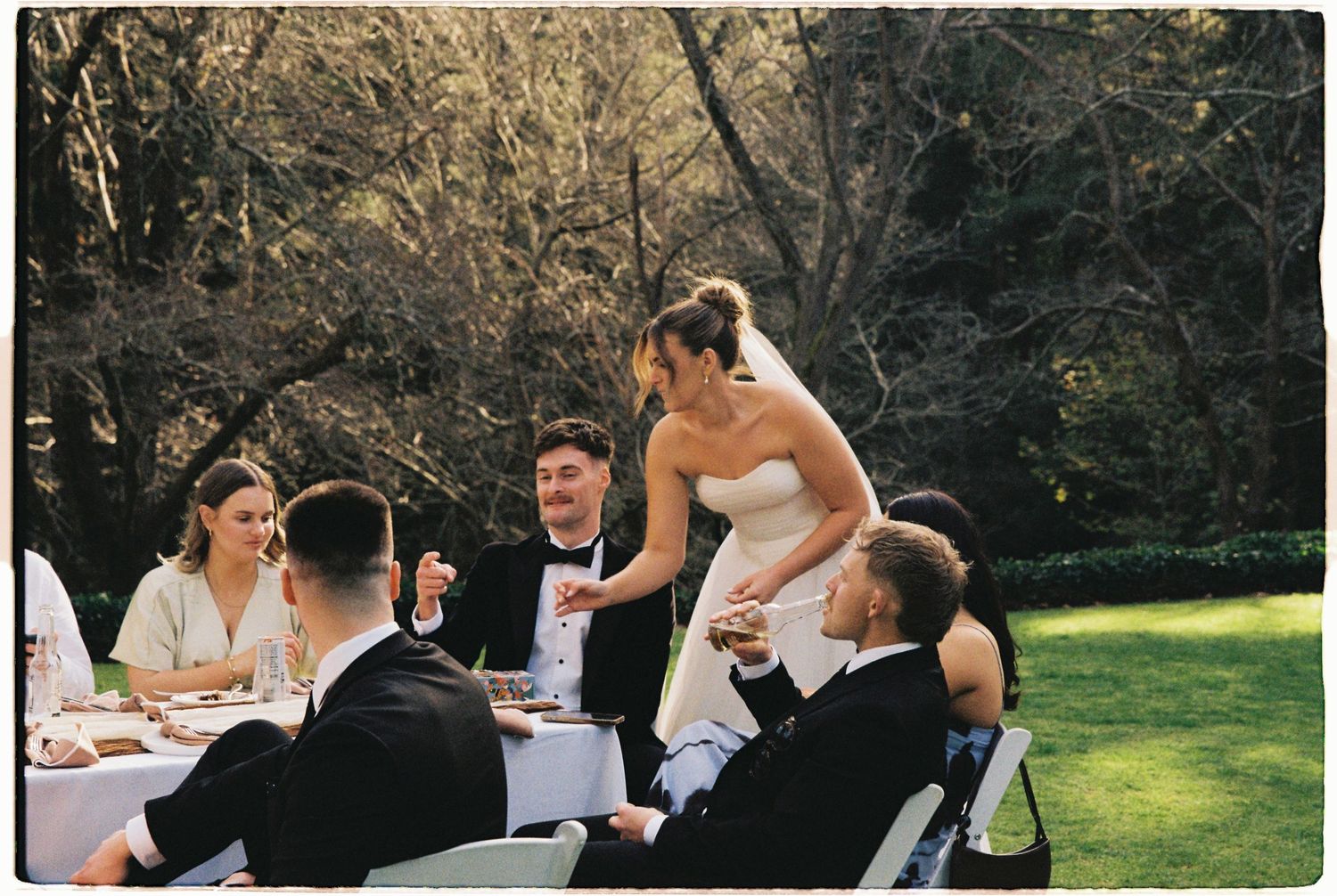 Bride in white dress leaning over wedding guests seated at outdoor reception table.