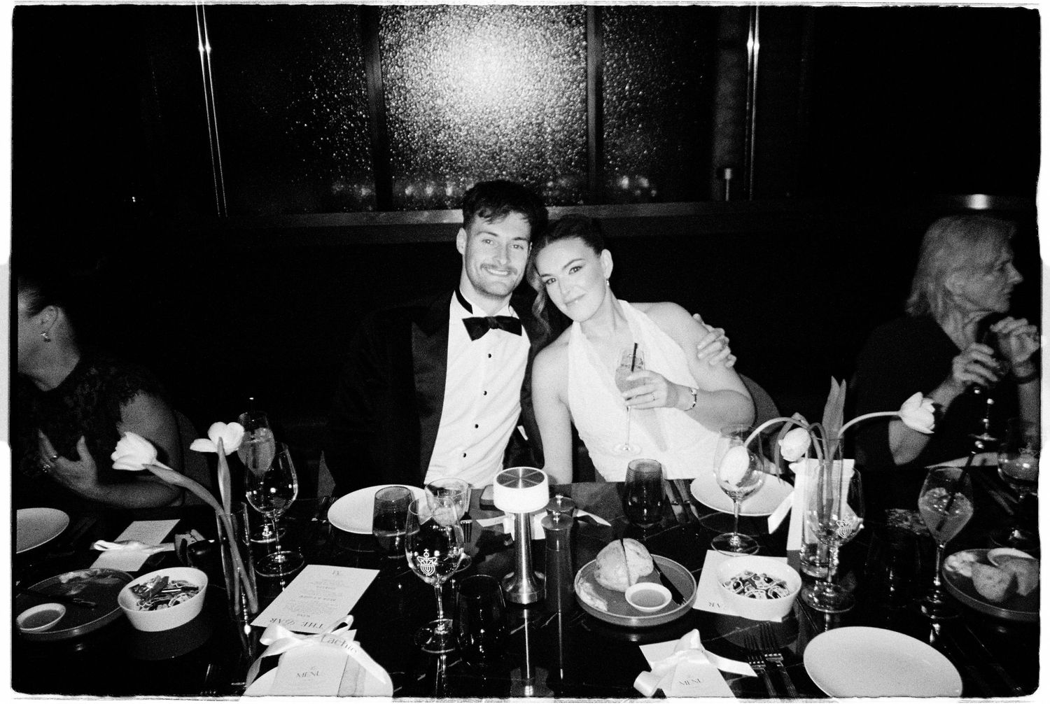 Black and white photo of a couple in formal attire smiling together at an elegantly set wedding reception dinner table.