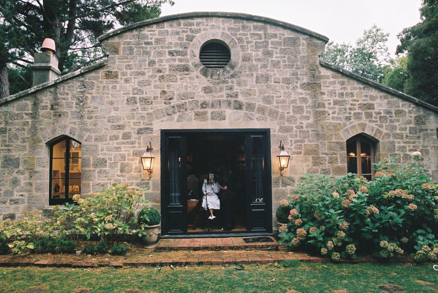 Stone chapel venue exterior with arched doorway, lanterns, and hydrangea bushes at dusk.