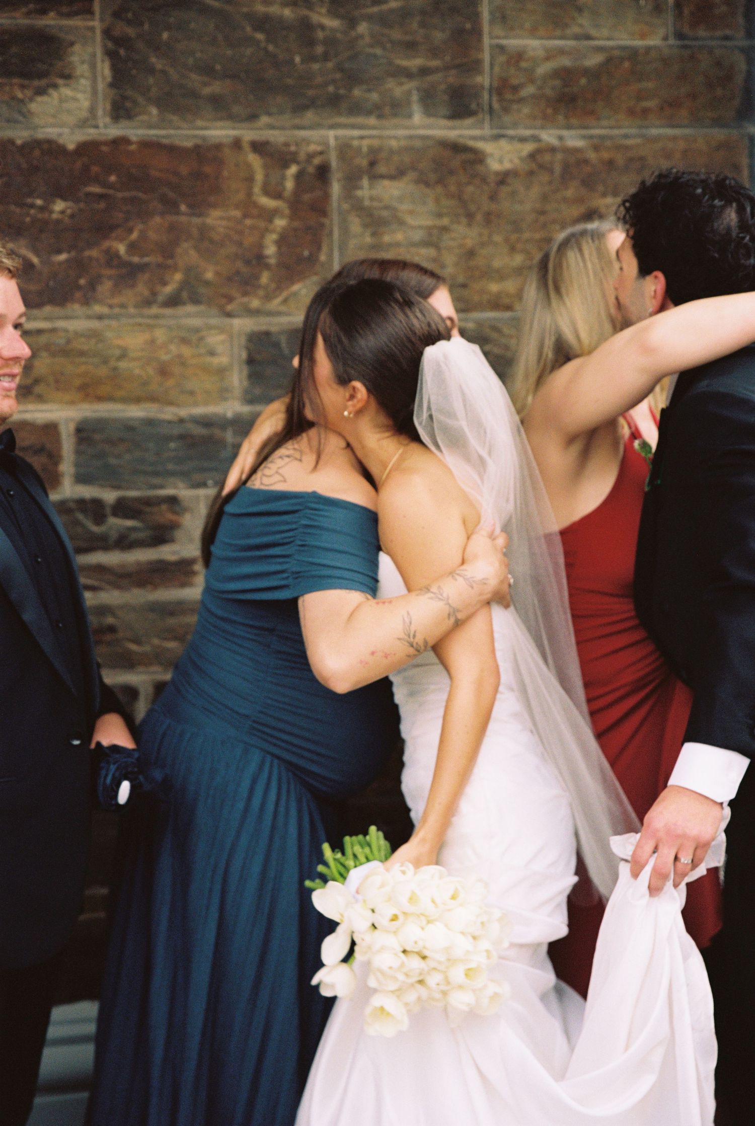 Bride in white gown embraces bridesmaid in teal dress outside stone church wall.