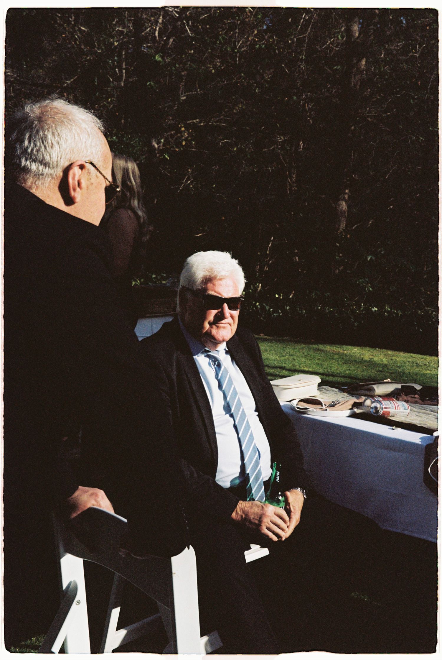 Elderly man in black suit and sunglasses sits outdoors holding a green bottle at a garden wedding reception.
