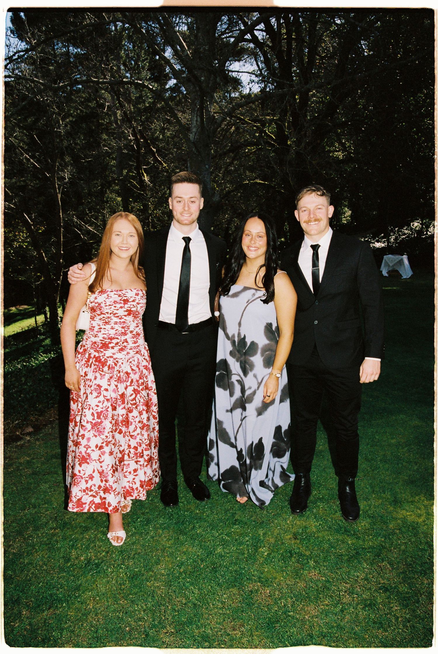 Four wedding guests in formal attire posing together on green lawn surrounded by tall trees.