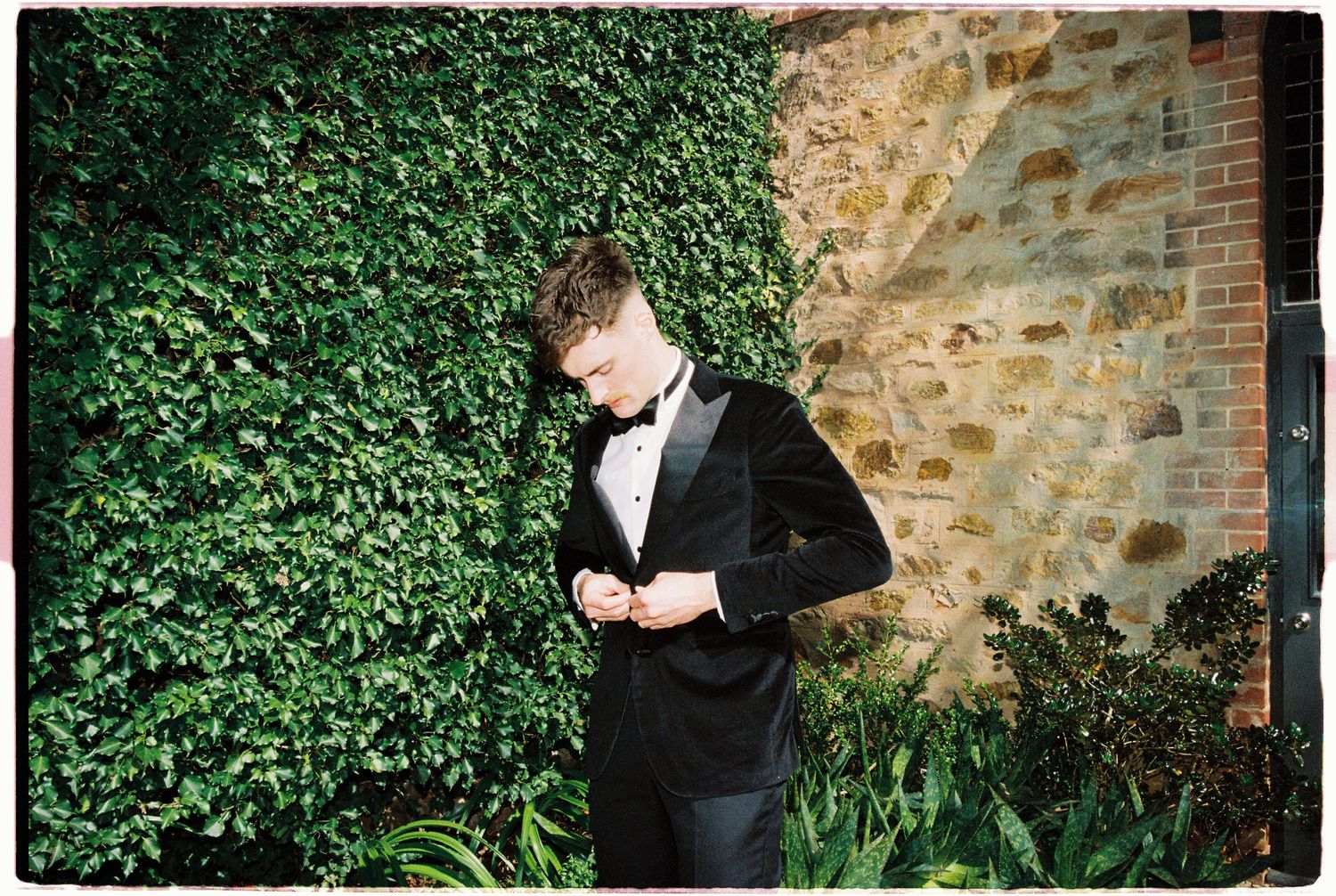 Groom in black velvet tuxedo adjusting jacket beside a stone wall covered in green ivy.
