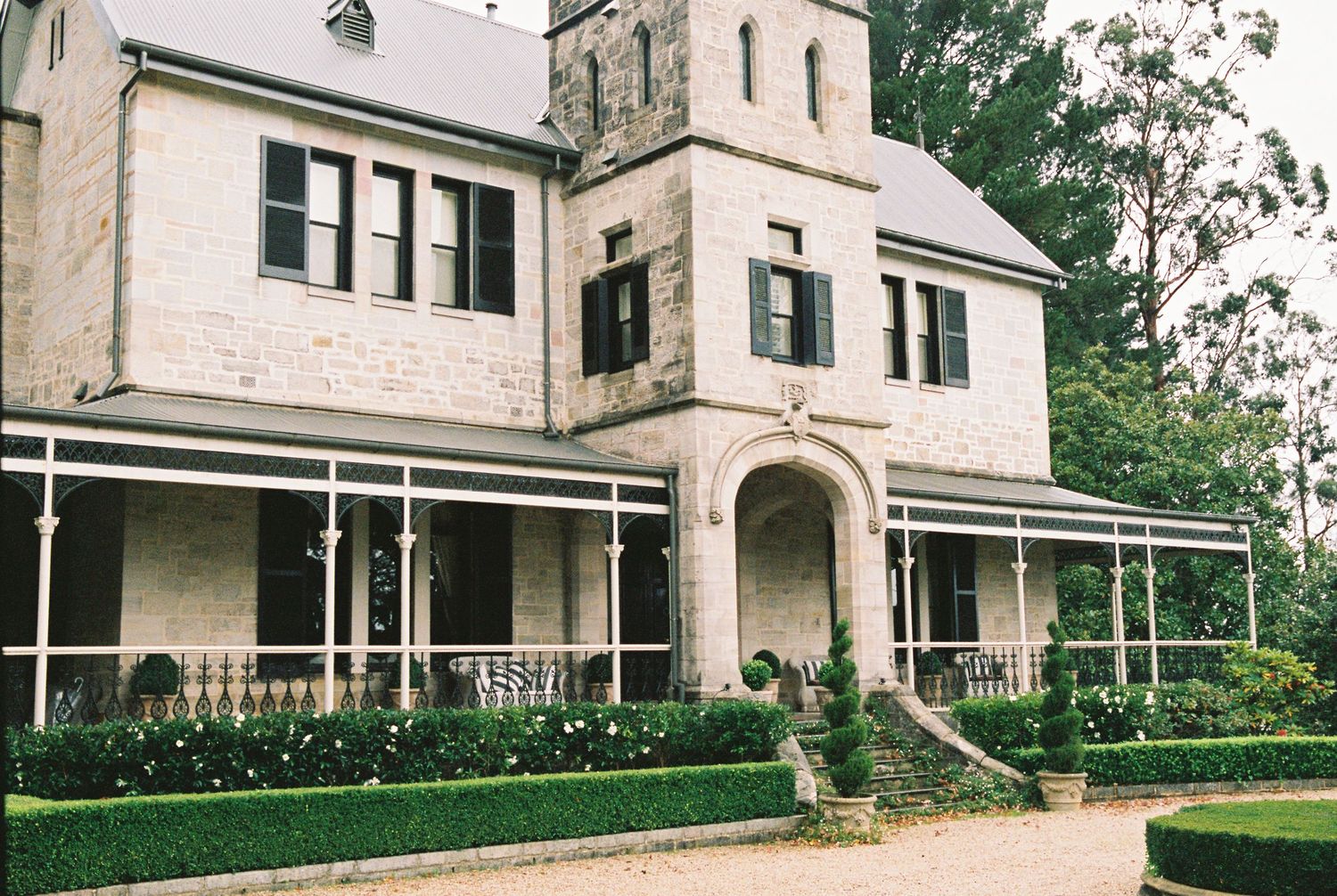 Elegant stone manor house with wraparound veranda and manicured hedges.