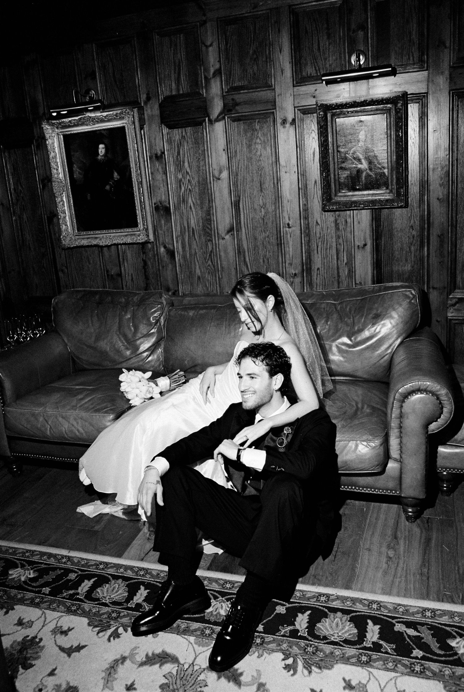 Groom sitting on floor smiling as bride in veil leans over him affectionately.