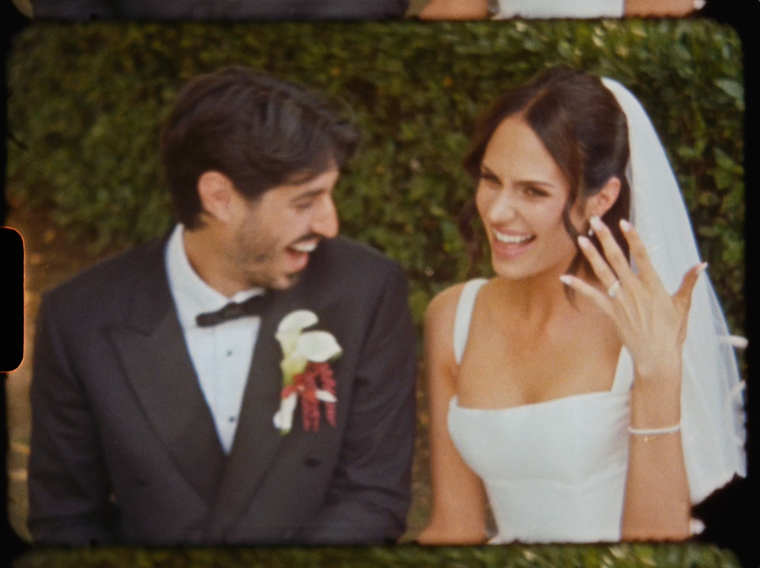 Smiling bride shows off her wedding ring while sitting beside her groom at their outdoor reception.