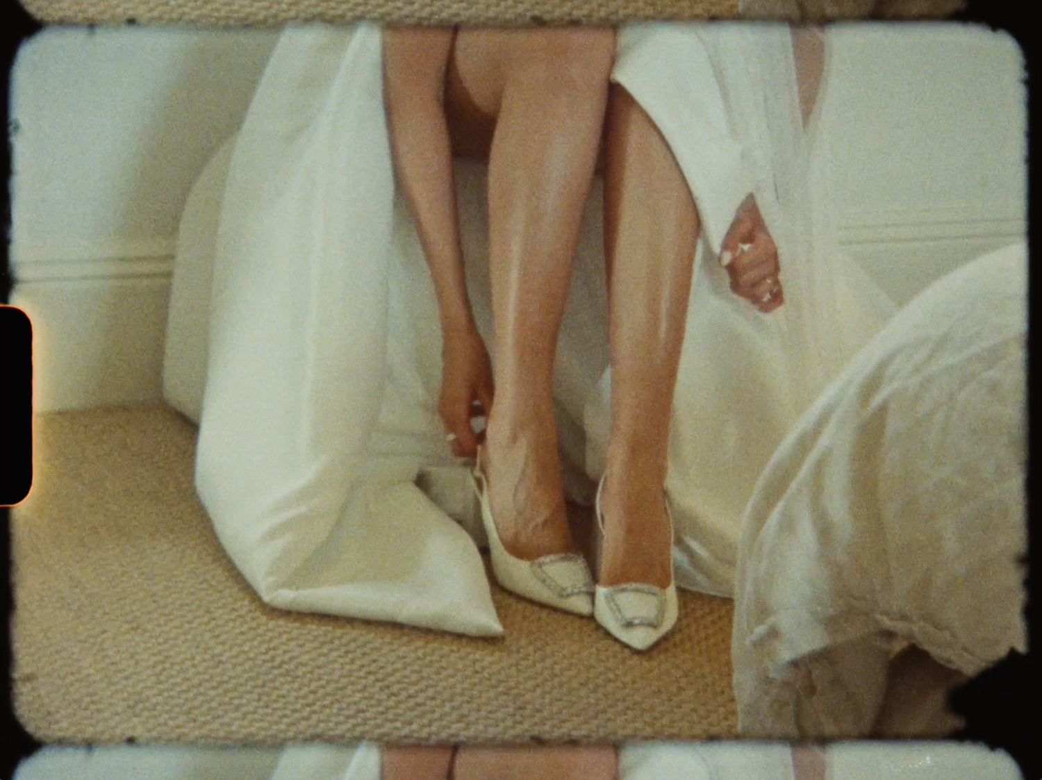 Film-style photo of a bride putting on white pointed-toe heels beneath her wedding gown.